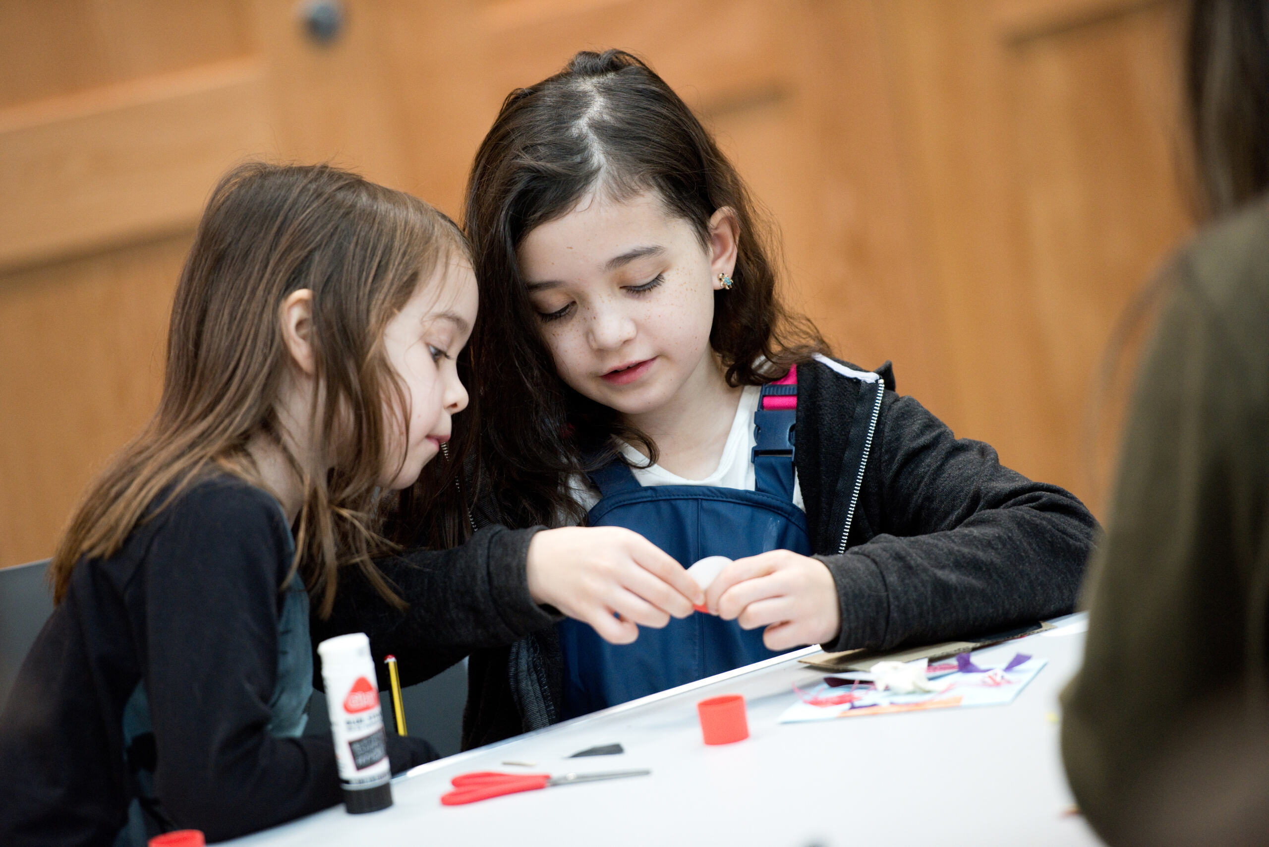 Hatton Gallery Two young girls sit at a table crafting together. One girl is watching what the other one is doing. There is glue and scissors on the table.