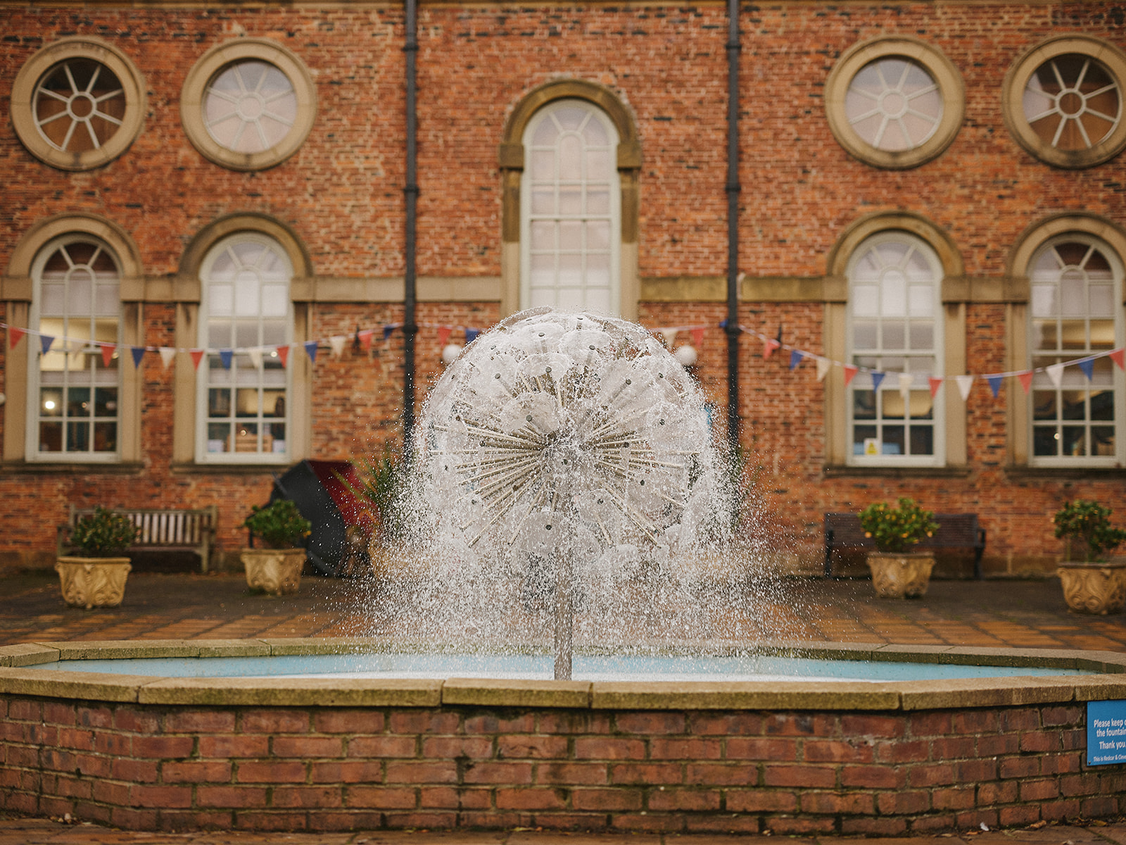 A fountain in front of a brick building that spins. As it is spins water is falling from the fountain.