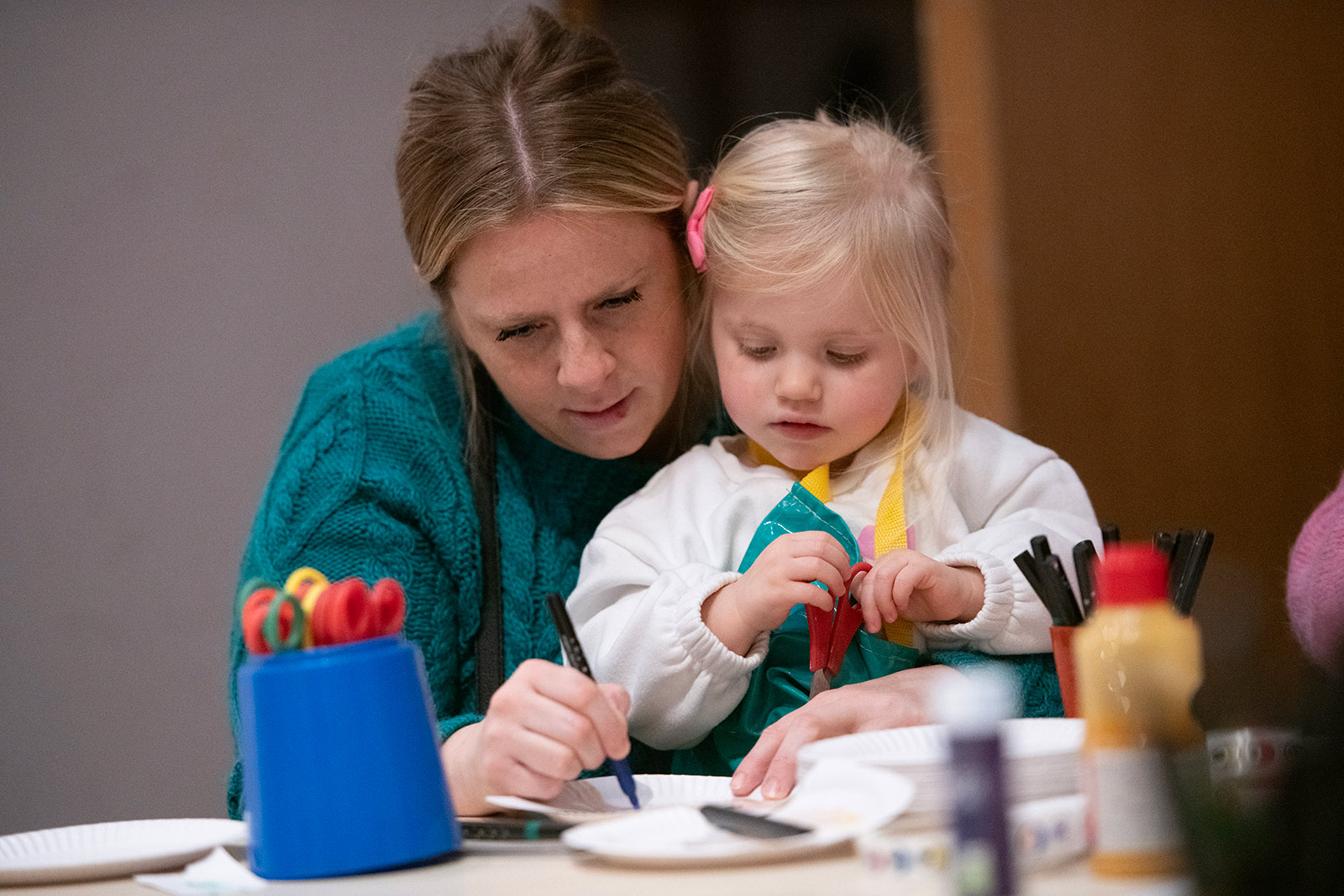 A woman sits at a craft table with her young daughter. The mum holds a black pen and the young girl holds red scissors.