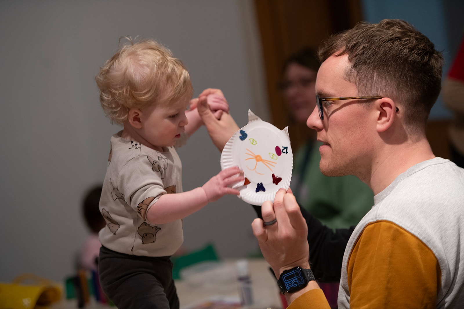 A father holds his young son's hand while in the other they hold up a cat face crafted out of a paper plate.