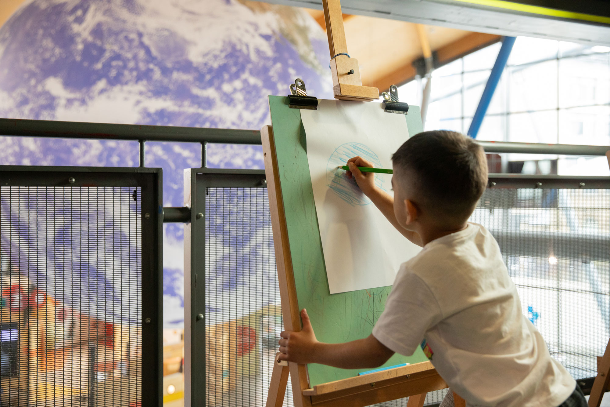 A boy draws planet Earth on a easel, copying a large replica that hangs from the ceiling in front of him.