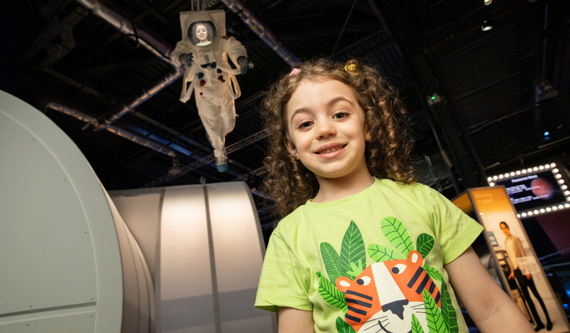 A girl stands in the foreground of the image. Behind her is an astronaut with her face on the mask.