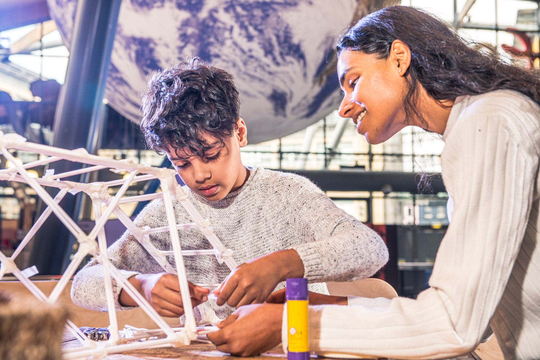 A woman sits with a young boy as he carefully constructs a bridge from white paper.