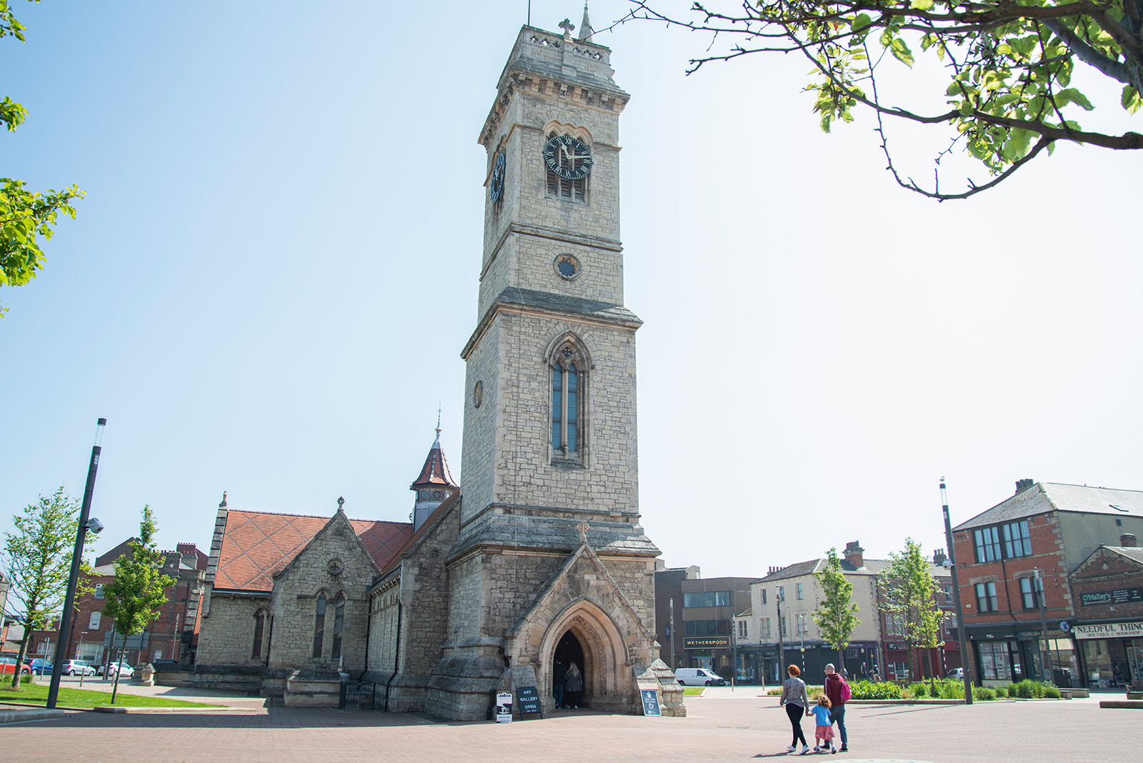A family walk up to a church with a tall clock tower in a town setting.