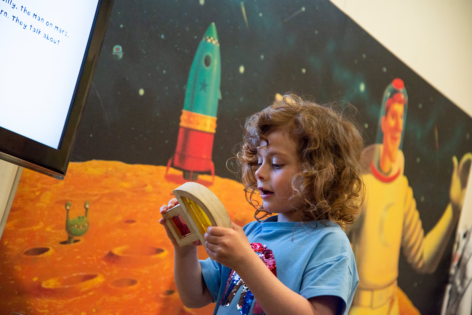 A young child holds a wooden timer in front of a wall painted with planets, aliens, a rocket and a spaceman.