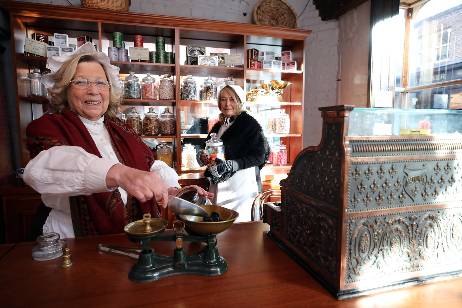 Two women dressed in Victorian dress and working in an old fashioned sweet shop, measuring out some sweets.