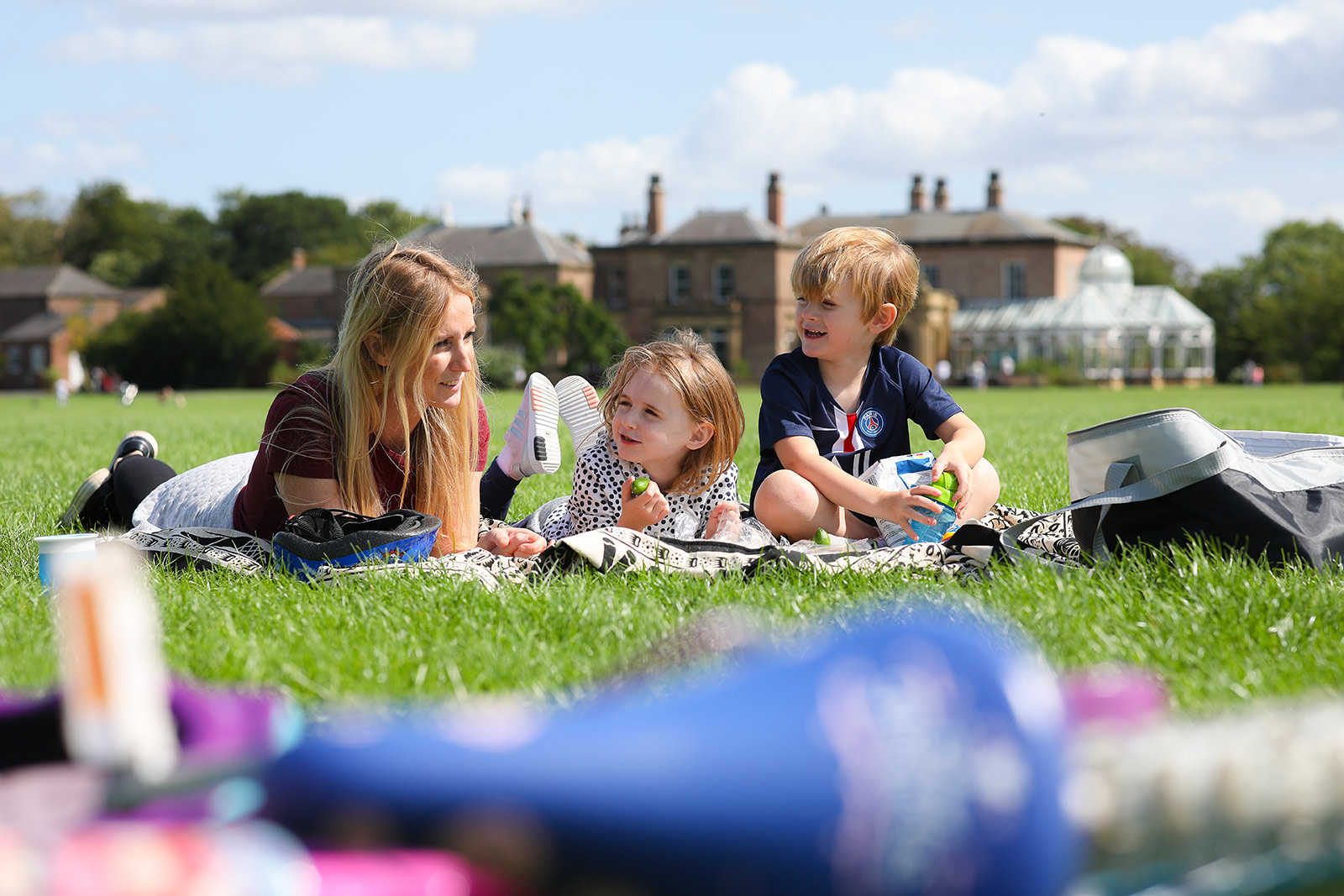 A mother and two young children sit on a picnic blanket on a grassy lawn in front of a large stone building.