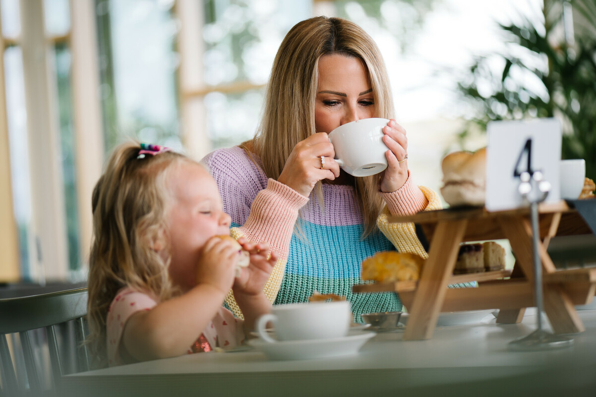 A mother and daughter sit at a table drinking from white mugs. A table number 4 is displayed and they are eating afternoon tea.