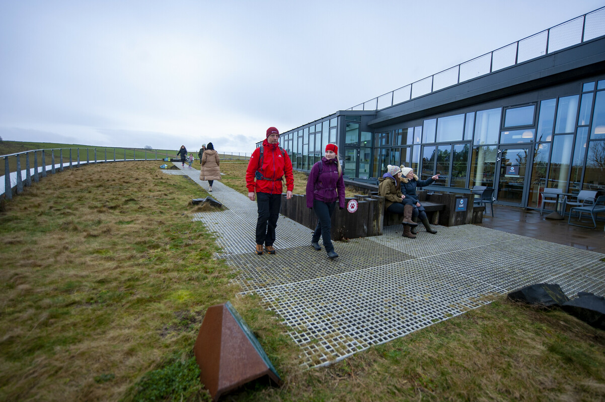 A man and teenage girl walk along a metal grated path in front of glass building. They are wearing waterproofs and hats.
