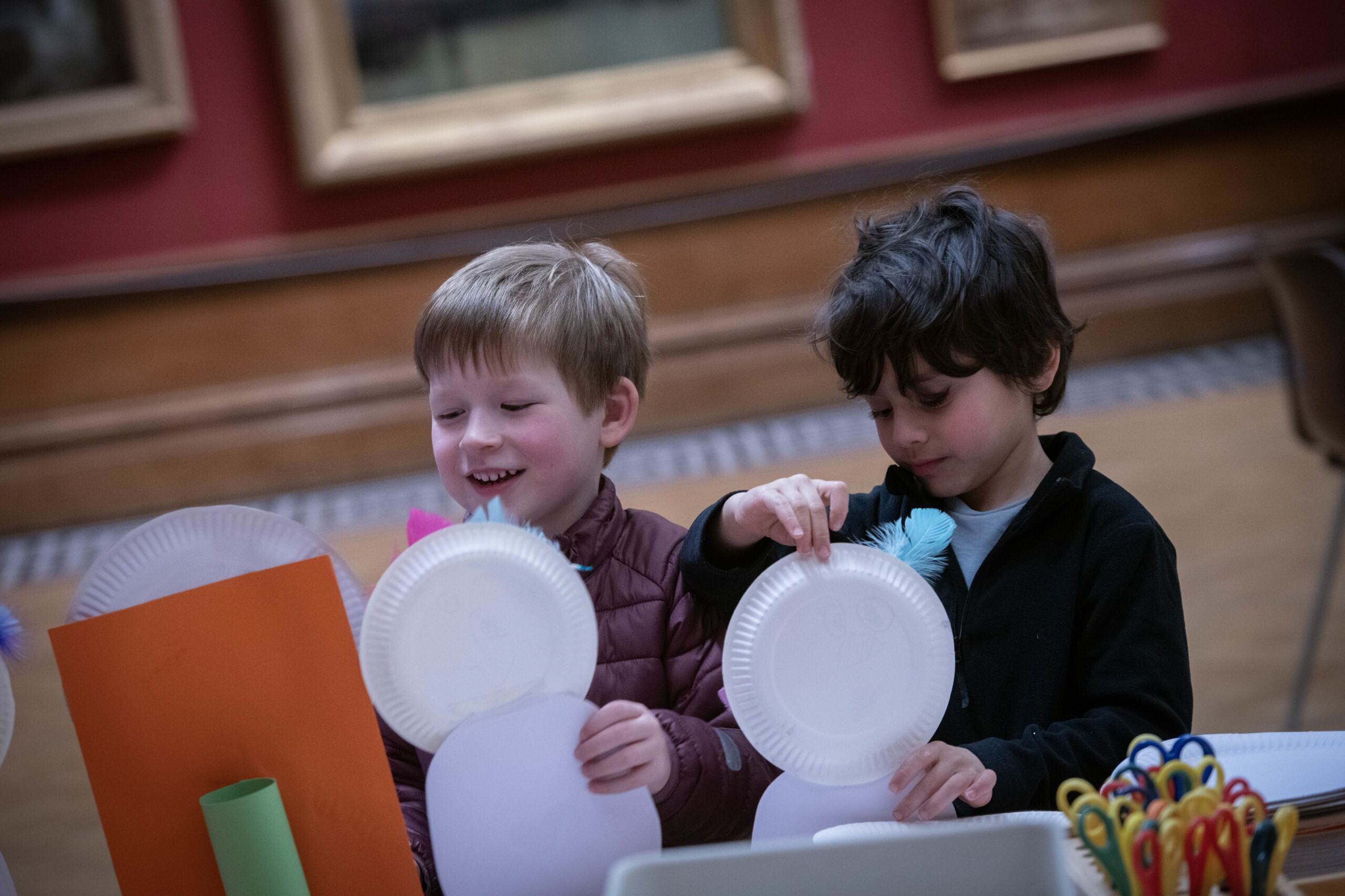 Two young boys sit at a craft table together holding white paper plates, one on top of the other.