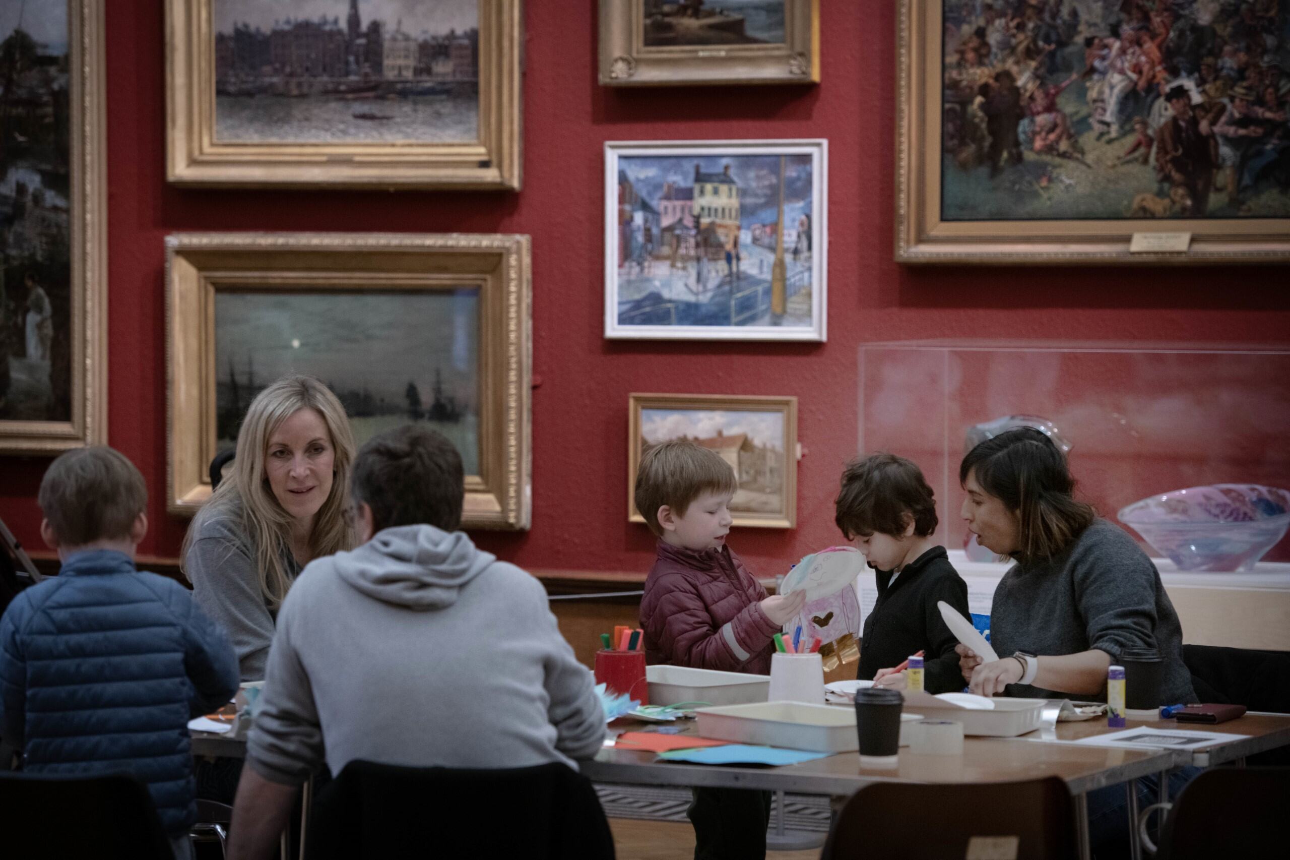 A group of both adults and children sit a craft table in a room with red walls and art work in frames.