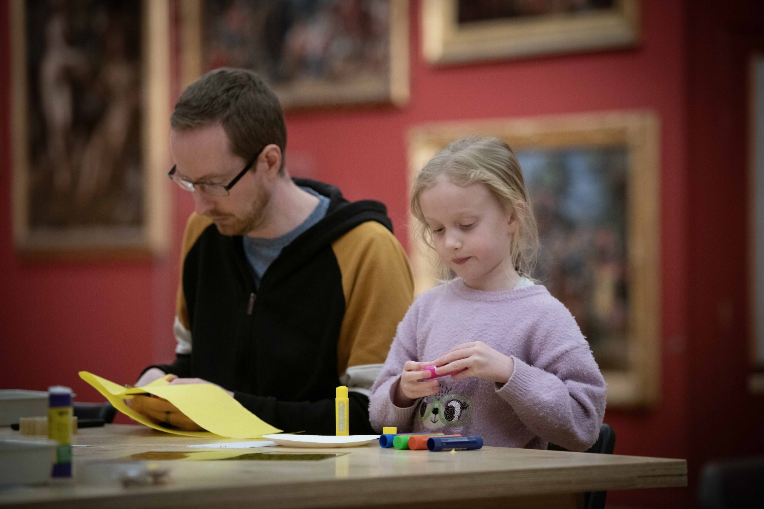 A father sits with his daughter at a craft table in a room with red walls and framed art work.