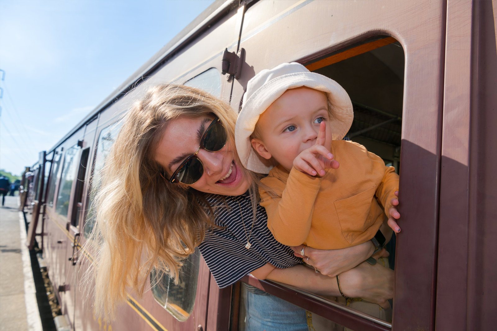 A mother holding a toddler hang out of the window of an old fashioned train carriage.