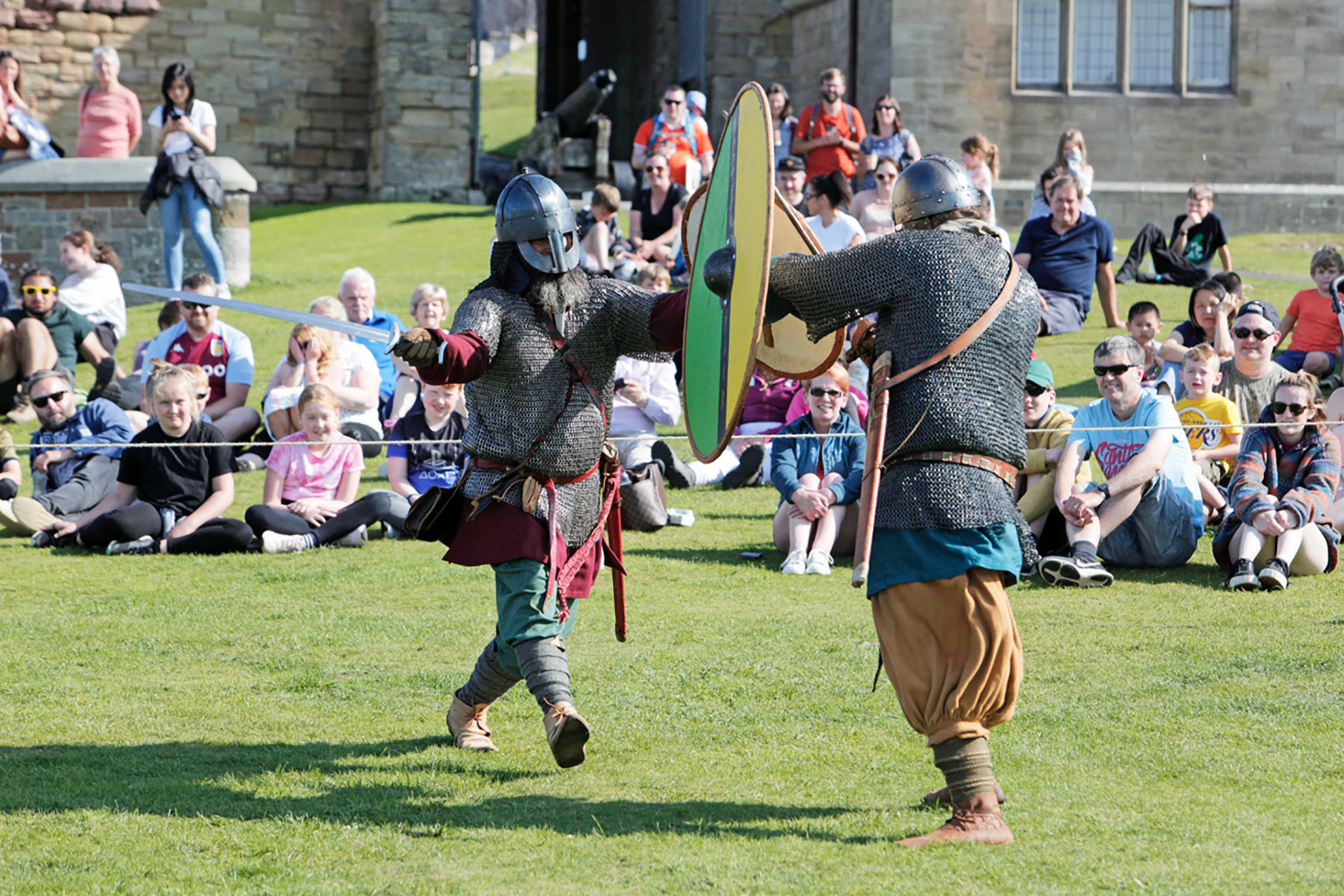 Two men dressed as vikings battle in front of a crowd who are sitting on a grassy bank.