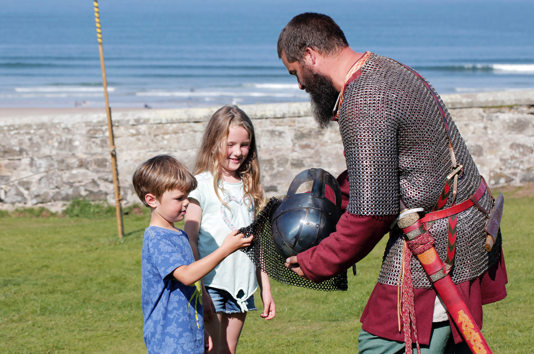 Two children look at a viking helmet held by a man dressed up as a viking.