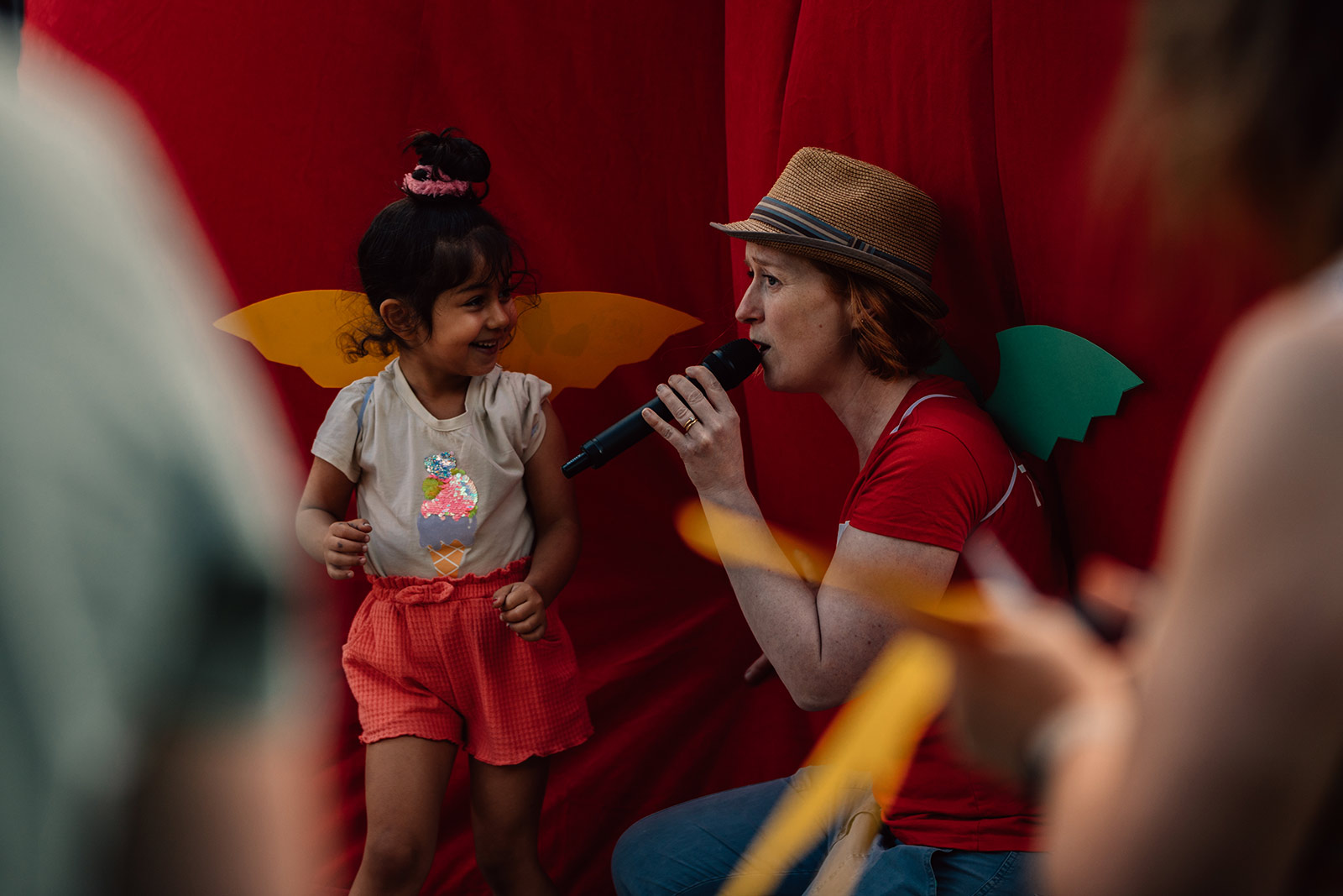 A woman wearing a brown bucket hat, red t-shirt and dragon wings, talks into a microphone next to a young girl also wearing dragon wings.