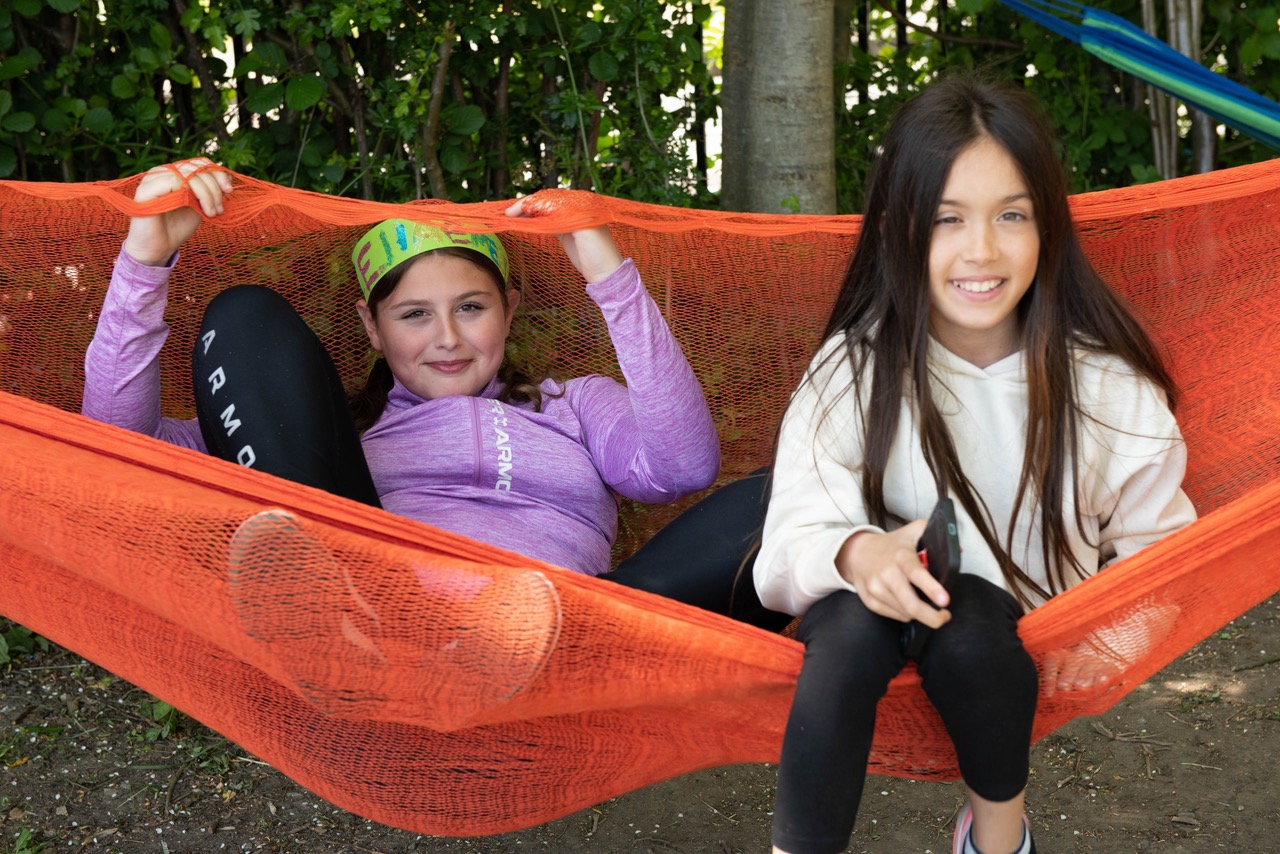 Two girls sit in an orange hammock tide between trees.