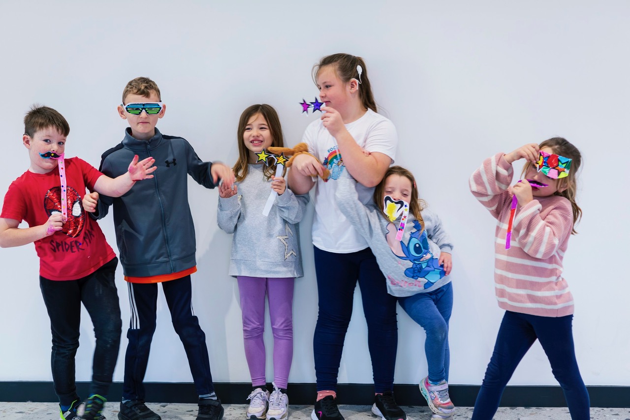 A group of six children stand in a line with different masks and glasses they have made held up to their faces. They are stood in front of a white background.