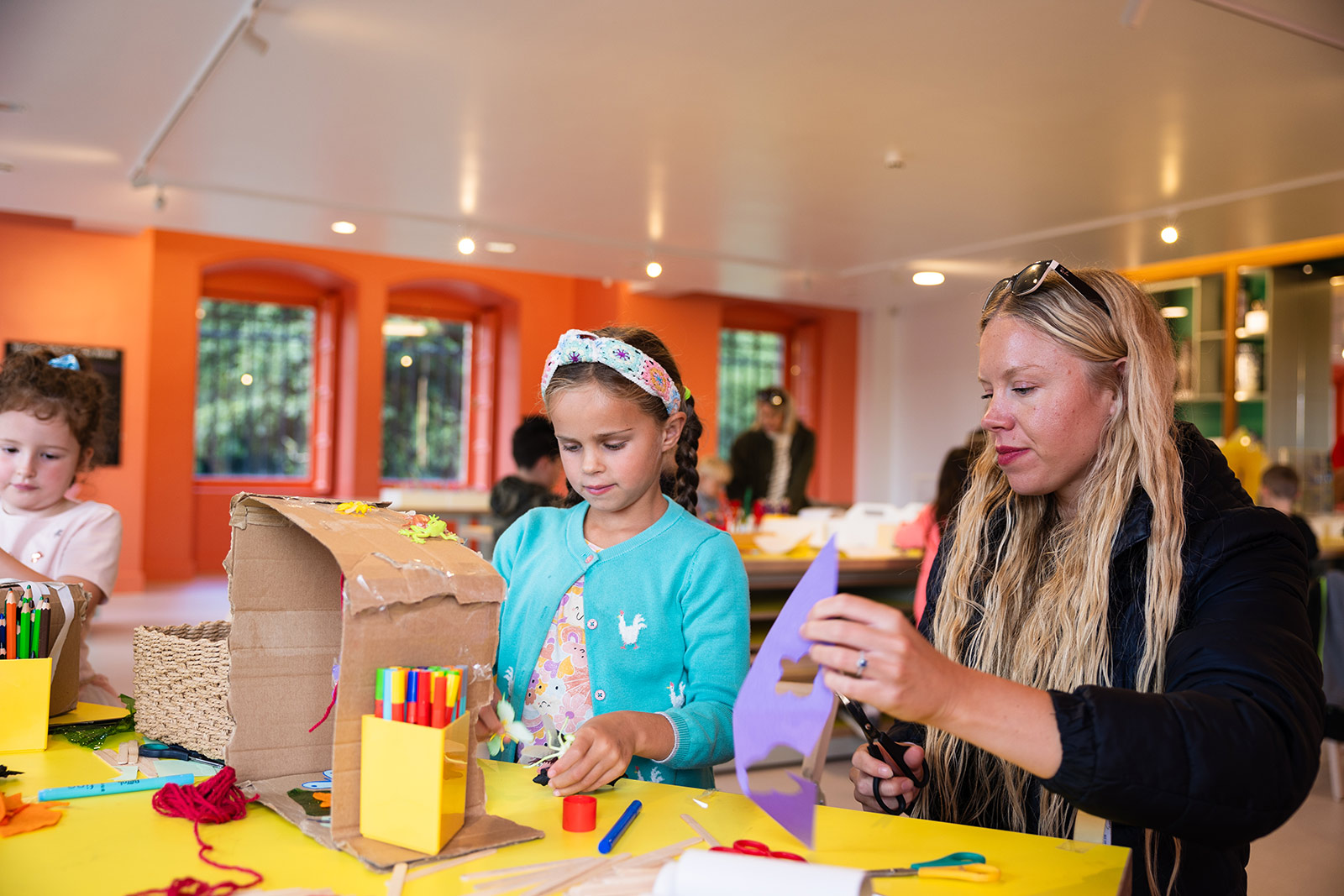 Two young girls sit at a craft table with their mother. They are making creations with cardboard and coloured paper.