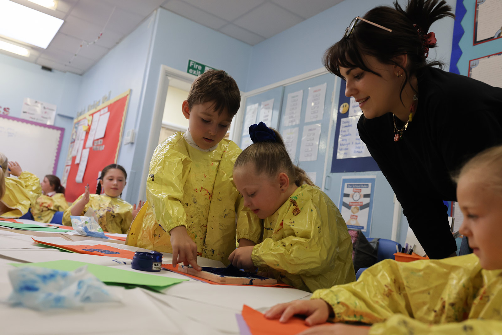 Children wearing yellow painting aprons with long sleeves sit around a craft table and using ink to make prints.