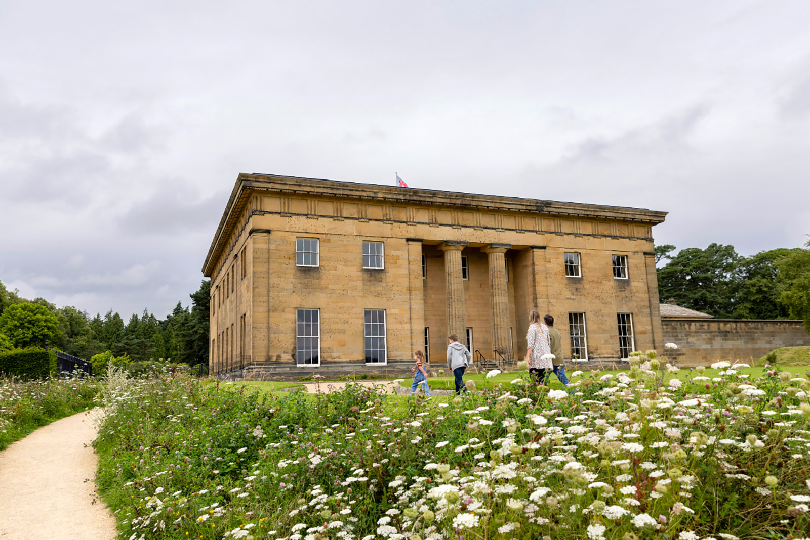 An image of the exterior of a grand hall set in formal gardens. A family group are walking in front of it.