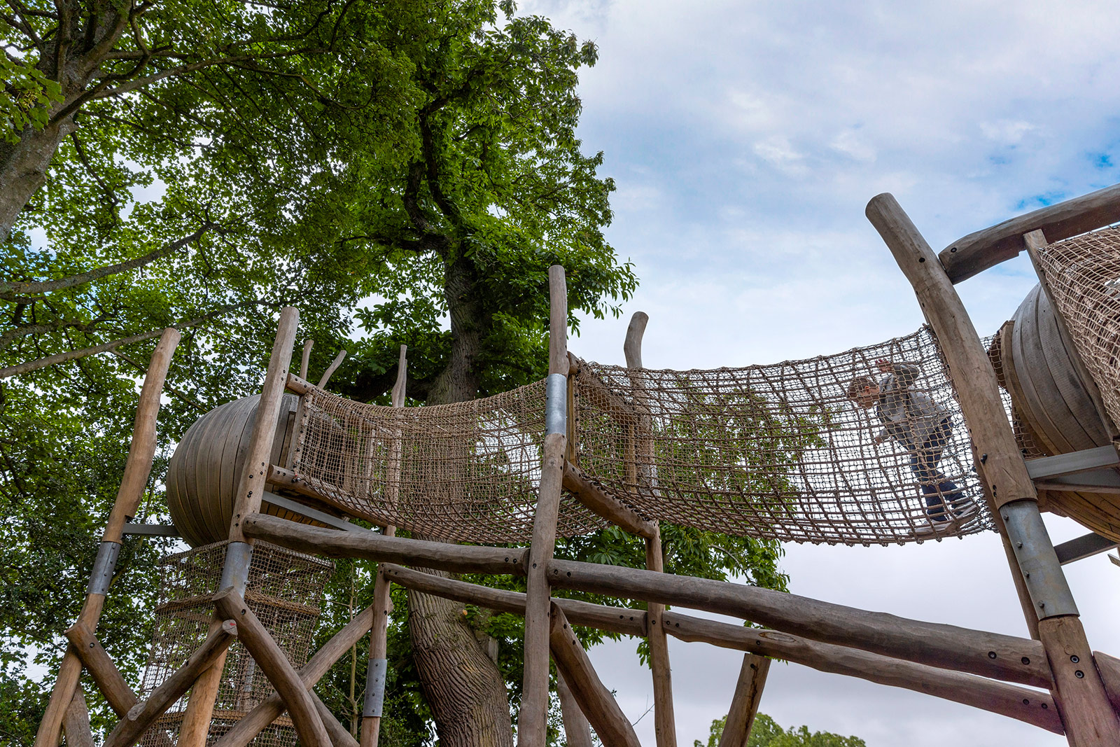 An image of a playground with ropes and treehouses. A child is walking through an enclosed rope tunnel.