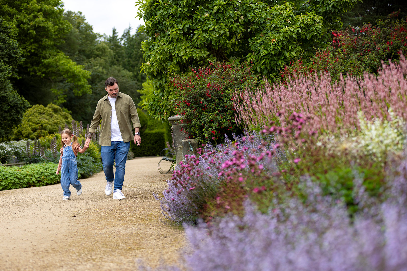A father and daughter walk hand in hand through formal gardens.