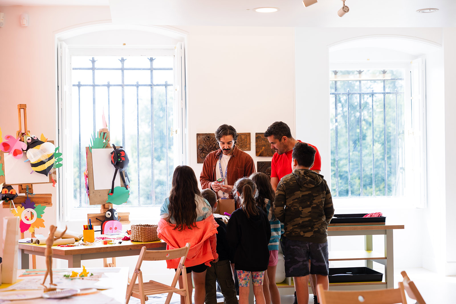 A man leads a children's activity in a light and airy room. He is surrounded by a group of children with an adult. There are easels with handmade bees on.