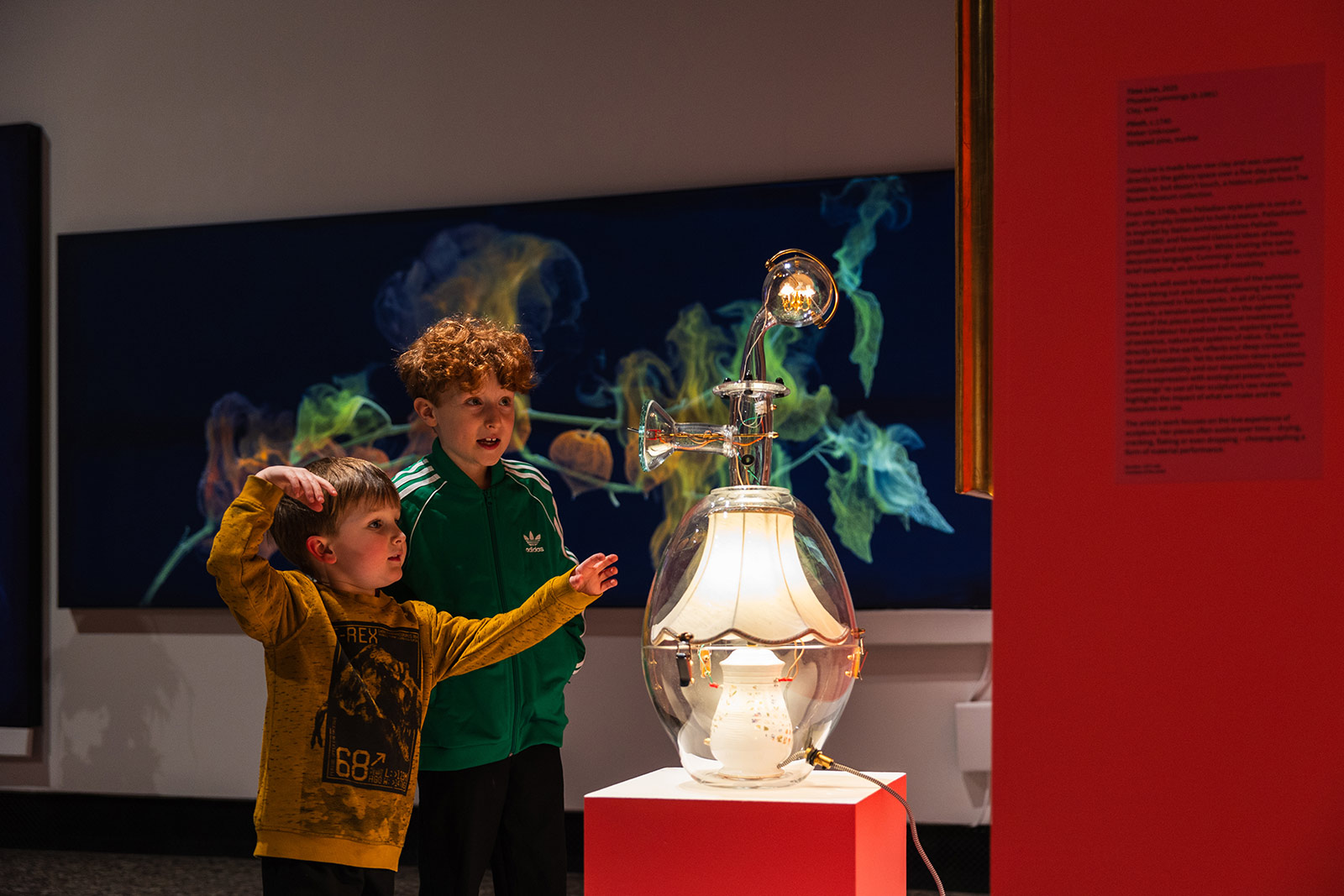 Two young boys looks at a lamp with wires and a lightbulb on top like a science experiment in a glass dome.