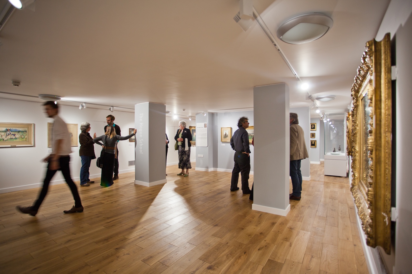 People wander around a gallery with a low ceiling and art work on the walls.