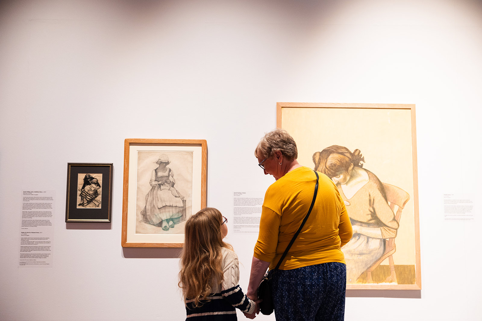 A grandmother holds a young girls and as they look at artwork on a white wall.