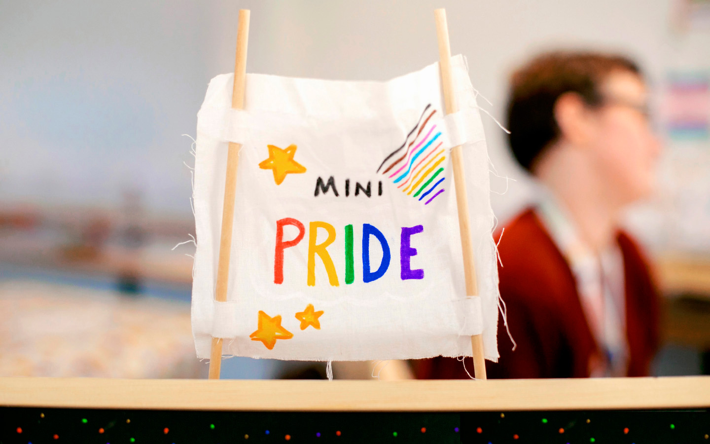 Mini Pride banner A mini pride banner made from white cloth and two wooden sticks stands on a table.