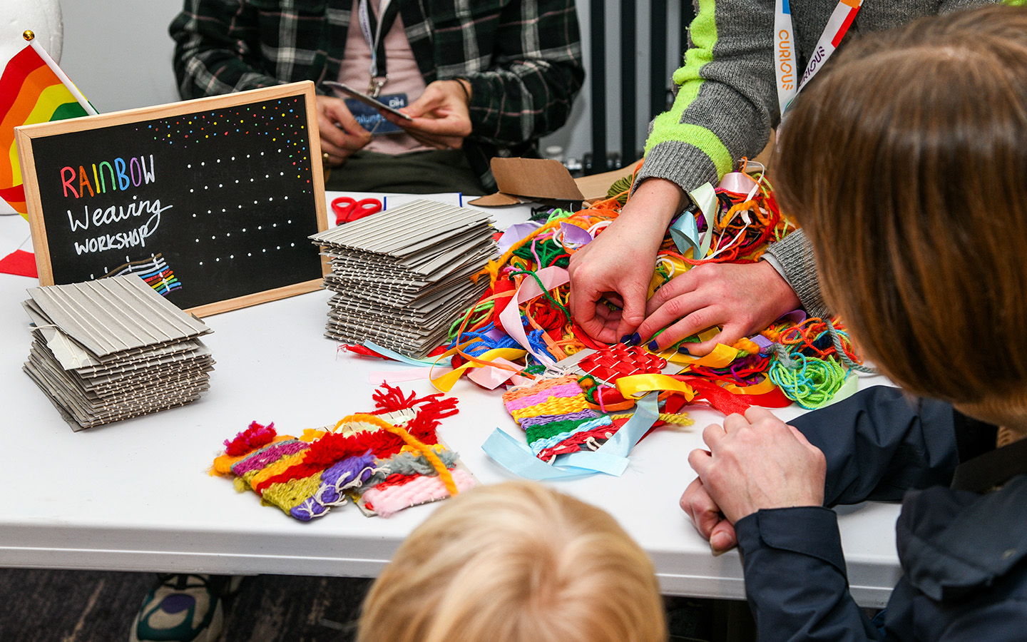 Mini Pride banner (Lauren Close) Colourful ribbons and craft materials are on a table with people stood around looking at the fabrics, only their hands are visible. A black chalk board reads Rainbow Wearing Workshop.