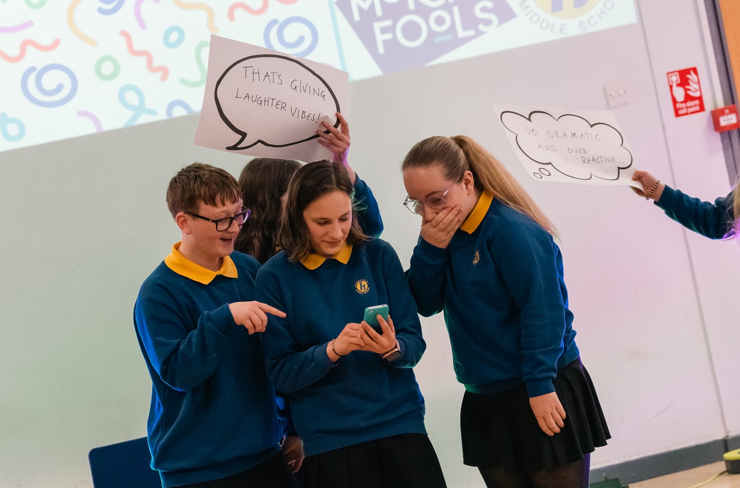 Three young people in school uniform crowd around a mobile phone, looking surprised at what's on it. Two you people behing them are holding up speech bubbles.