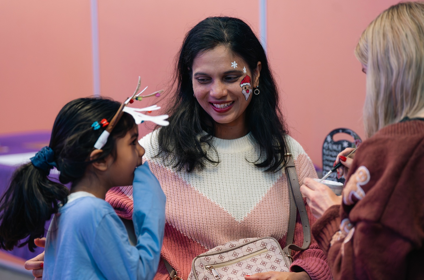 An adult smiles down at a child. They have a Santa sticker on their face and the child is wearing reindeer ears.