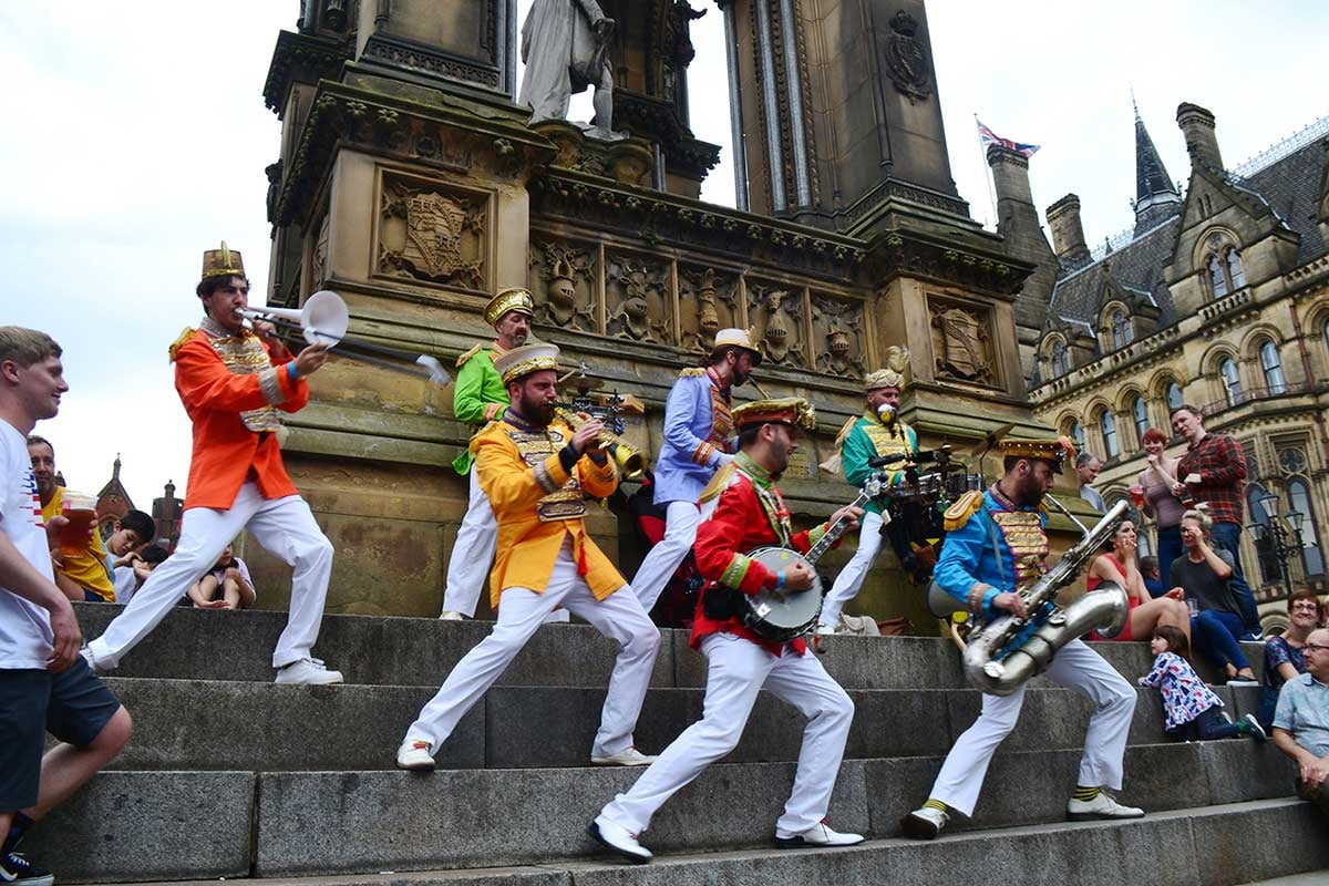 A band stand on the steps of a monument weating brightly coloured jackets and playing a range of instruments.