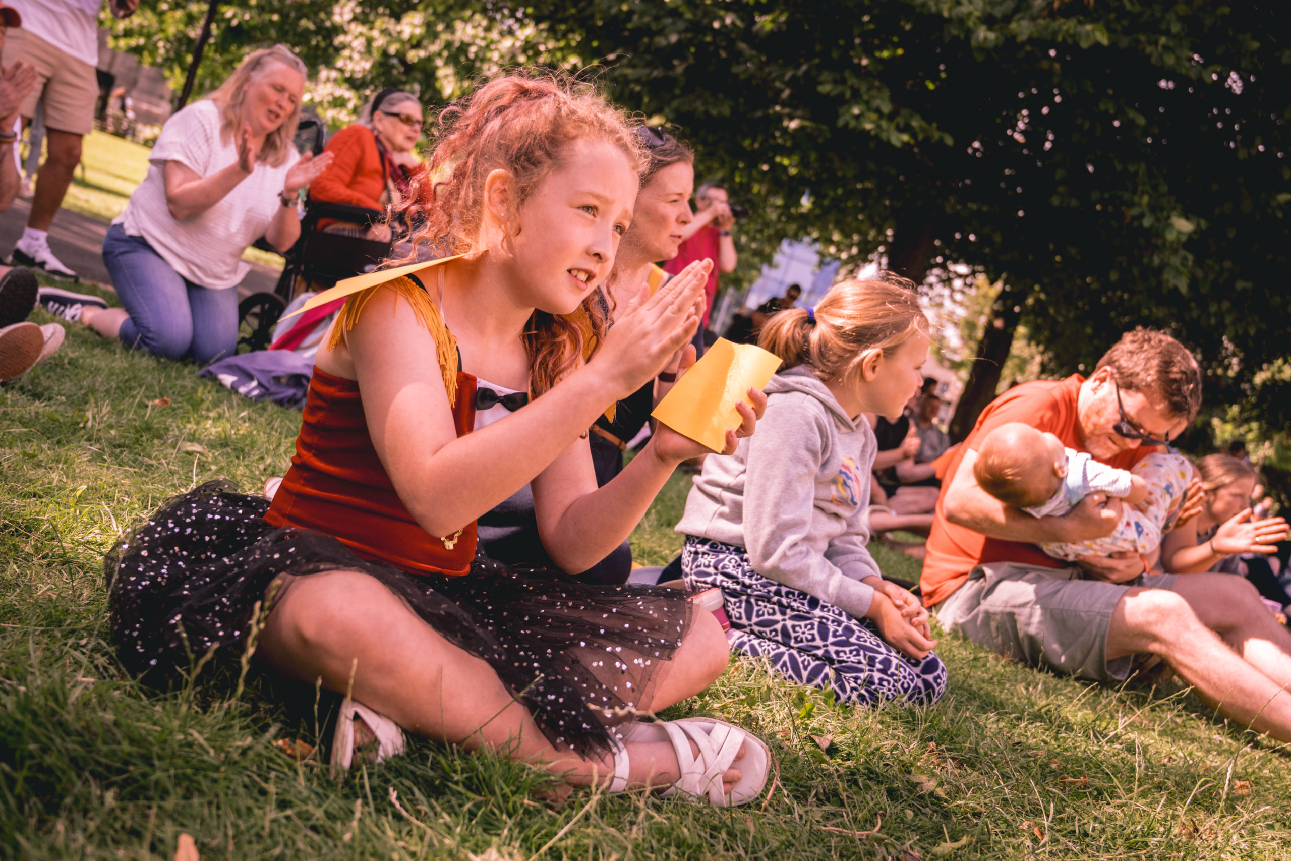 Lots of children sit on the grass watching something and clapping.