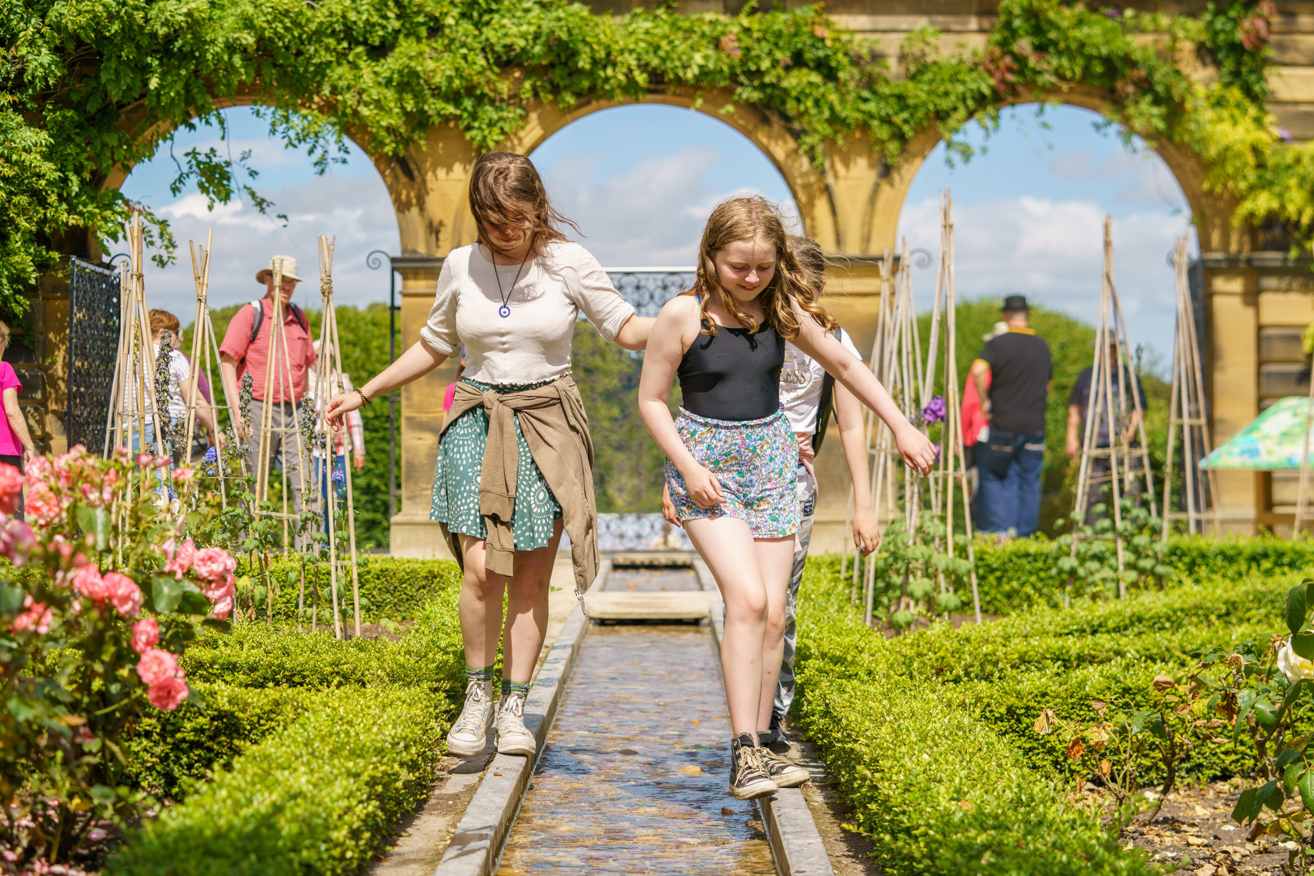 two older children balance on curbs either side of a stream of water. There are large stone archways behind the, and flowers in bloom.