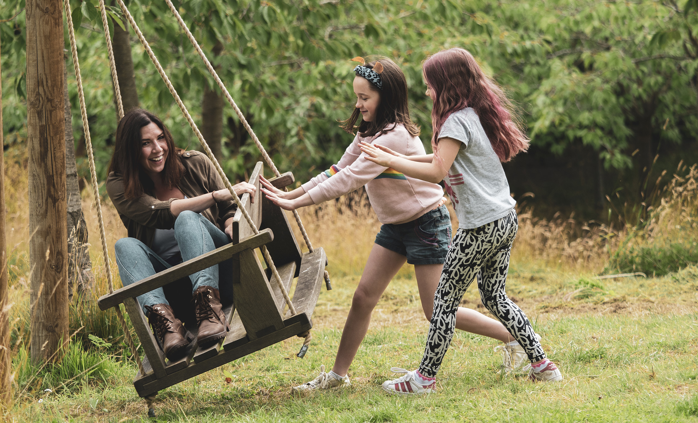 Two children push a laughing adult in a large wooden swing.