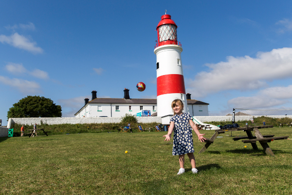 A young girl throws a ball up into the air as she stands in front of Souter Lighthouse on a grassy bank.