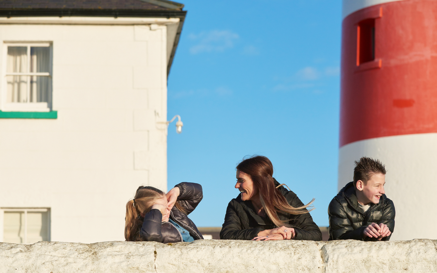 Three family members lean on a whitewashed wall with the lighthouse visible behind them