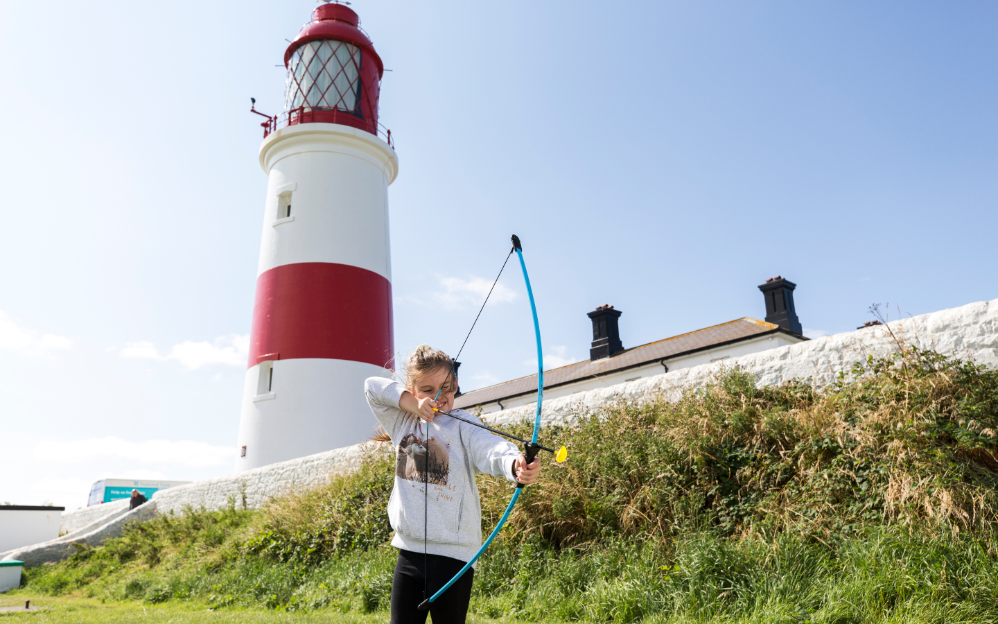 A child fires a toy bow and arrow with a lighthouse in the background.