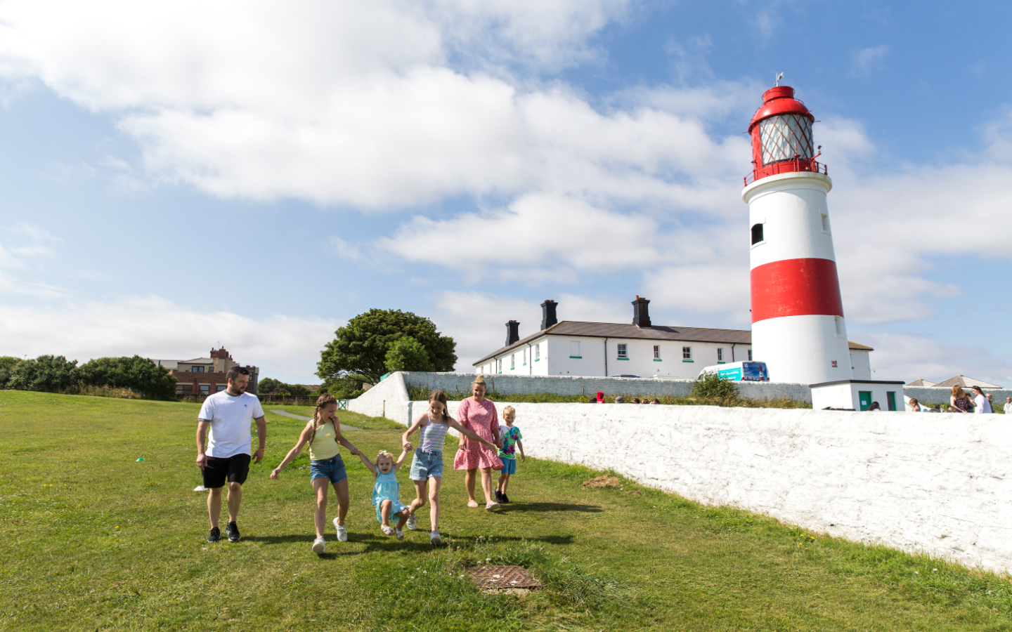 A family of six walk hand in hand with the lighthouse and white buildings on their right.