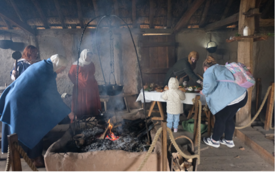 A scene inside an Anglo Saxon round house featuring re-enactors tending to a fire and family visitors.