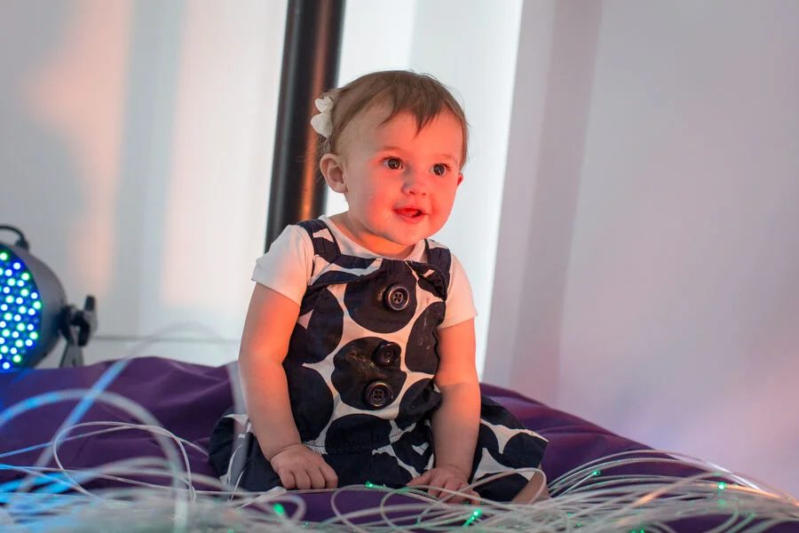 A toddler inside Baltic's sensory room smiles. They are sitting on a giant purple cushion surrounded by coloured fairy lights.