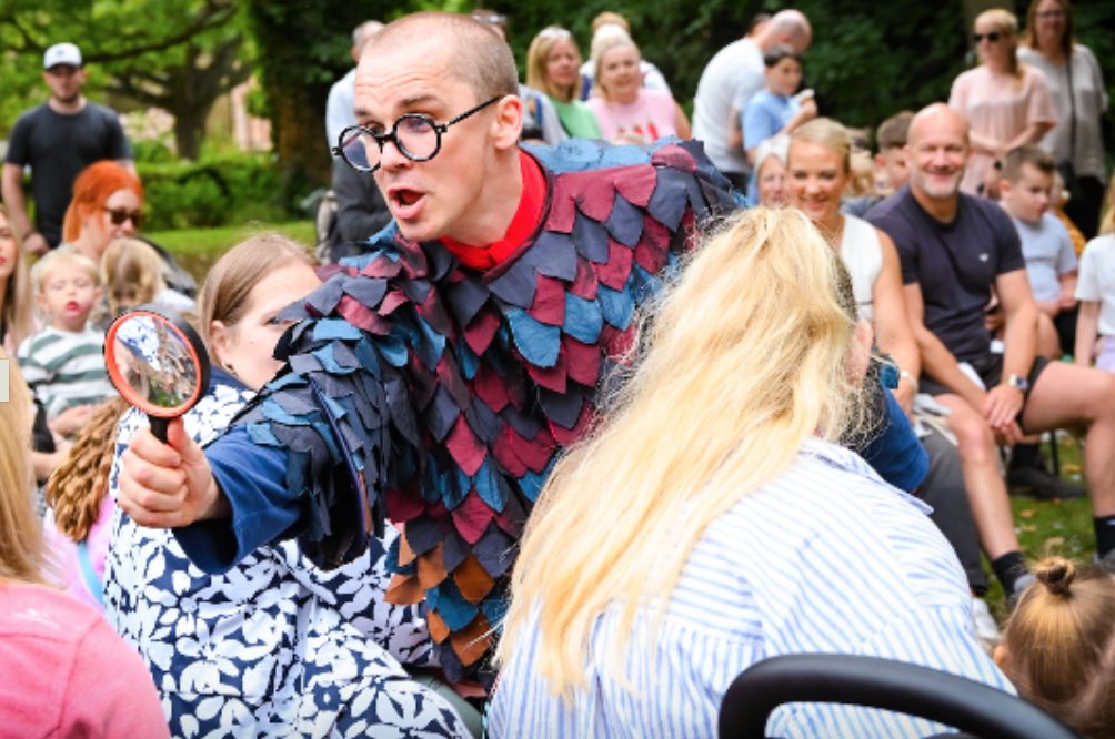 A man leans through a crowd of people, holding a large magnifying glass.