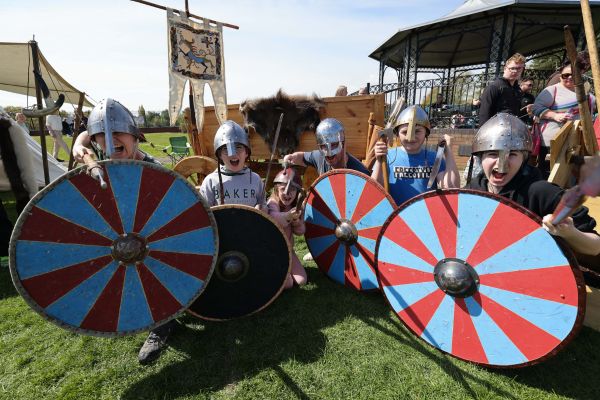 A group of young children are dressed as vikings and holding large red and blue shields in front of them.