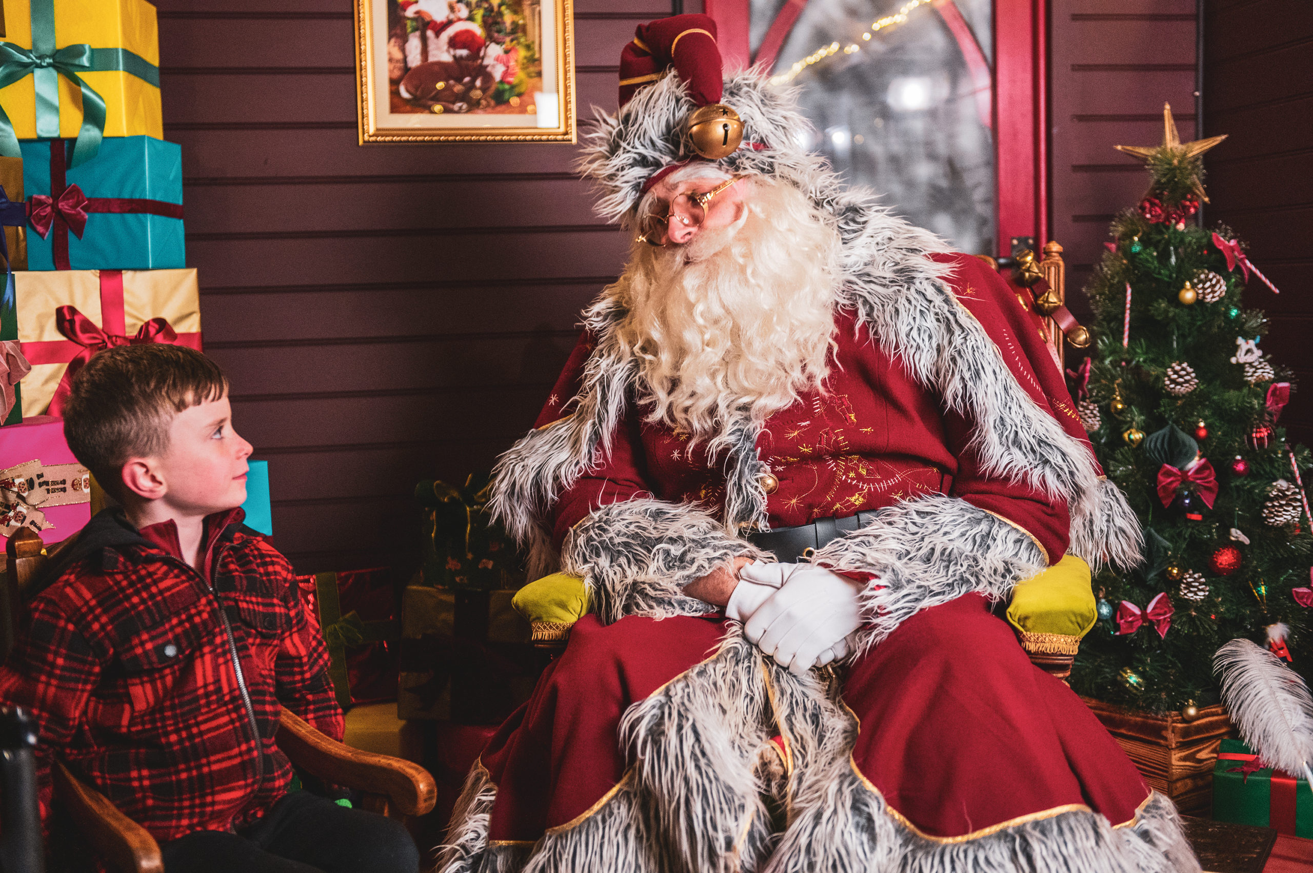 An image of a young boy wearing a red checked short, sitting next to Father Christmas with a stack of presents and a Christmas tree behind them.